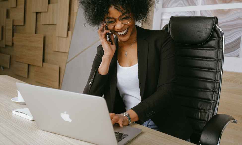 black woman on phone call happy, working on Macbook laptop